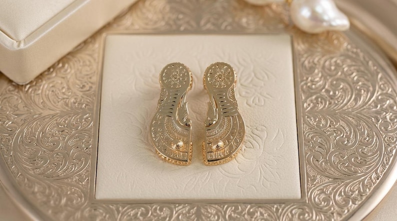 May include: A pair of gold-toned earrings featuring a flat, elongated shape with intricate, symmetrical engraved patterns. Each earring has a circular top section and a wider, curved bottom section, both decorated with fine lines and small dots.