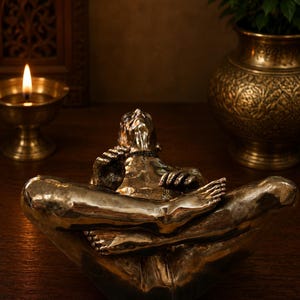 May include: A metallic bronze-toned sculpture of a figure in a seated meditative pose with legs crossed. The figure has a smooth, reflective surface, defined facial features, and hands resting on the knees, capturing a serene and focused posture.