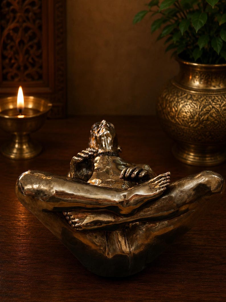 May include: A metallic bronze-toned sculpture of a figure in a seated meditative pose with legs crossed. The figure has a smooth, reflective surface, defined facial features, and hands resting on the knees, capturing a serene and focused posture.
