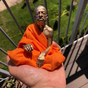 May include: A bronze-colored statue of a seated figure in orange robes. The statue has detailed facial features and is seated in a meditative pose. The figure's hands are visible, and the orange fabric drapes around the figure.