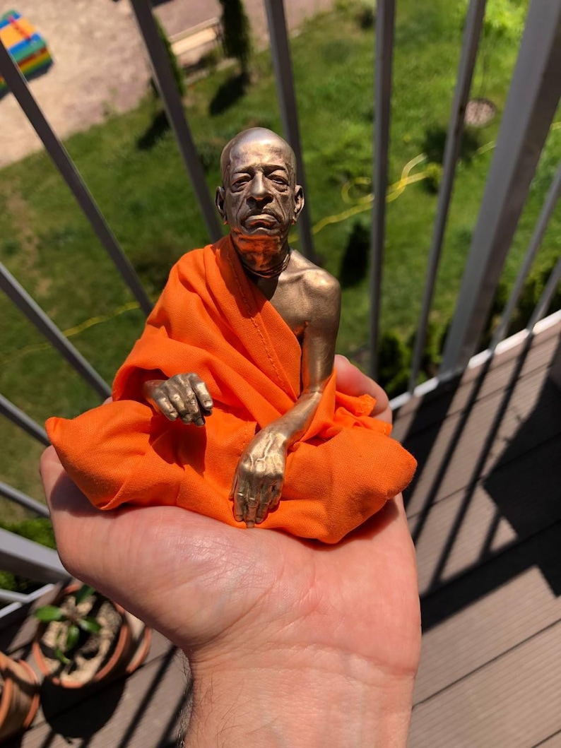 May include: A bronze-colored statue of a seated figure in orange robes. The statue has detailed facial features and is seated in a meditative pose. The figure's hands are visible, and the orange fabric drapes around the figure.