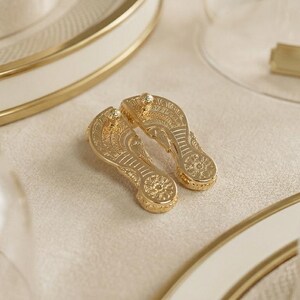 May include: A pair of gold-toned metal flip-flop shaped charms featuring intricate embossed patterns on the surface. Each charm has a textured sole design with a circular detail at the heel and a thong strap structure, reflecting a metallic finish.