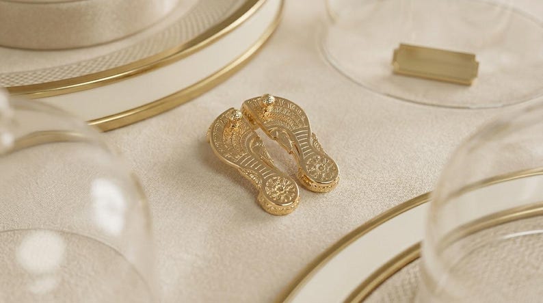 May include: A pair of gold-toned metal flip-flop shaped charms featuring intricate embossed patterns on the surface. Each charm has a textured sole design with a circular detail at the heel and a thong strap structure, reflecting a metallic finish.