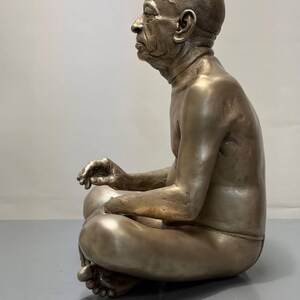 May include: A metallic bronze-toned sculpture of a seated figure in a cross-legged position. The figure is depicted in profile, showing a bald head, facial features, and a bare torso. The sculpture has a smooth, reflective finish throughout.