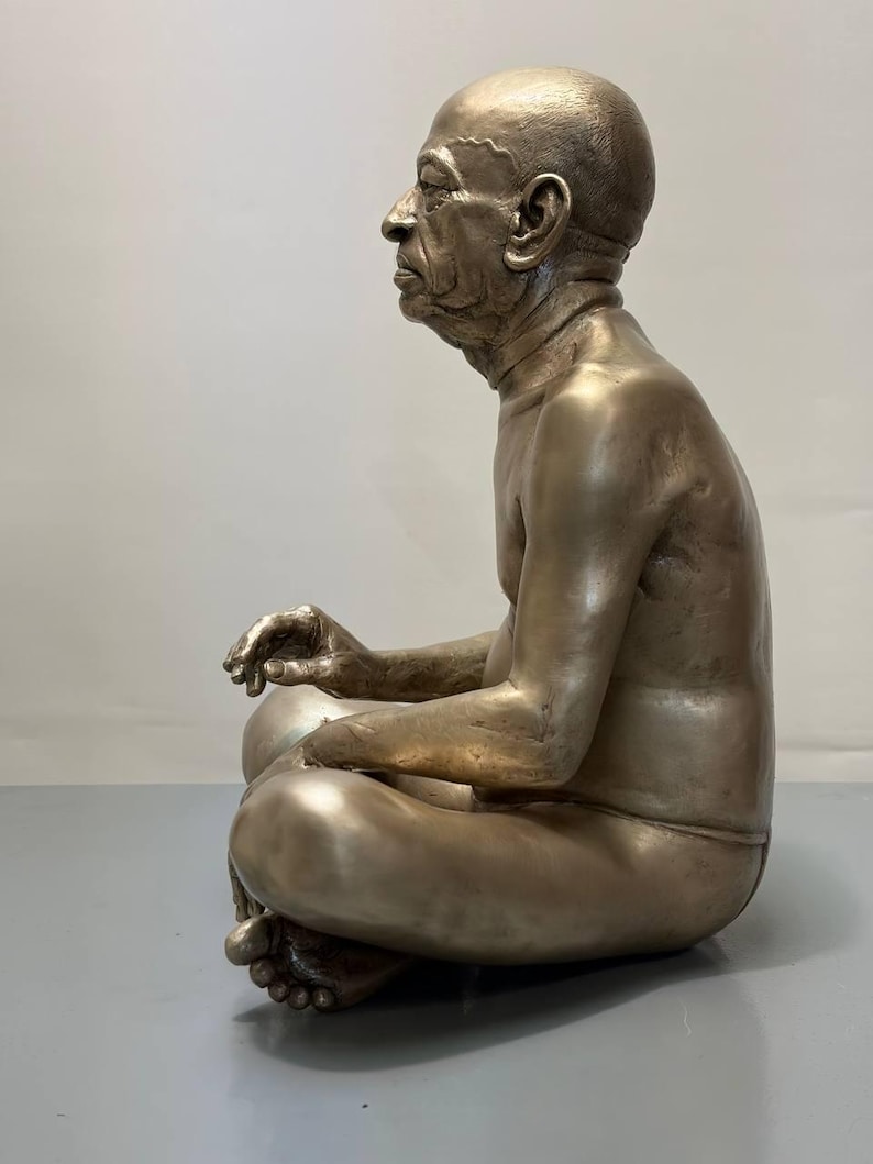 May include: A metallic bronze-toned sculpture of a seated figure in a cross-legged position. The figure is depicted in profile, showing a bald head, facial features, and a bare torso. The sculpture has a smooth, reflective finish throughout.