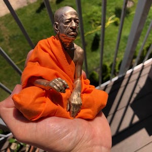 May include: A bronze-colored statue of a seated figure in orange robes. The figure has a bald head and is seated in a meditative pose. The statue is held in a person's hand, with a green lawn and metal railing in the background.