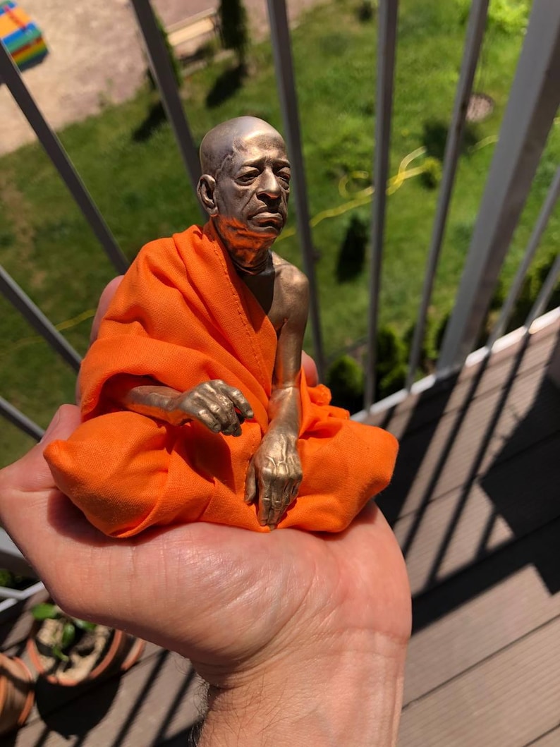 May include: A bronze-colored statue of a seated figure in orange robes. The figure has a bald head and is seated in a meditative pose. The statue is held in a person's hand, with a green lawn and metal railing in the background.