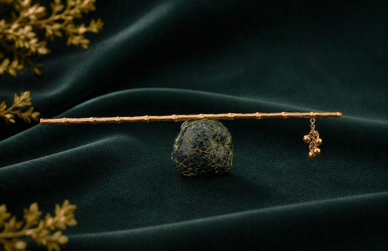 Puede incluir: Un palillo para el cabello en tono dorado dise&ntilde;ado para parecer un tallo de bamb&uacute;, con una superficie texturizada y nudos segmentados. Una peque&ntilde;a cadena dorada con tres diminutos dijes esf&eacute;ricos cuelga de un extremo del palillo sobre una piedra.