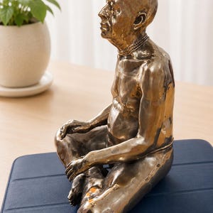 May include: A metallic bronze-toned sculpture of a seated figure in a meditative pose. The figure has a bald head, a defined facial profile, and a multi-layered necklace. The surface of the sculpture has a reflective, uneven texture throughout.