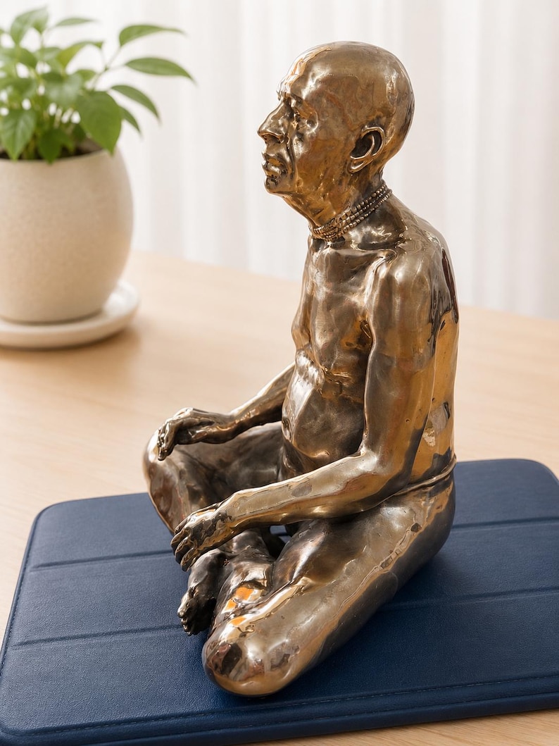May include: A metallic bronze-toned sculpture of a seated figure in a meditative pose. The figure has a bald head, a defined facial profile, and a multi-layered necklace. The surface of the sculpture has a reflective, uneven texture throughout.