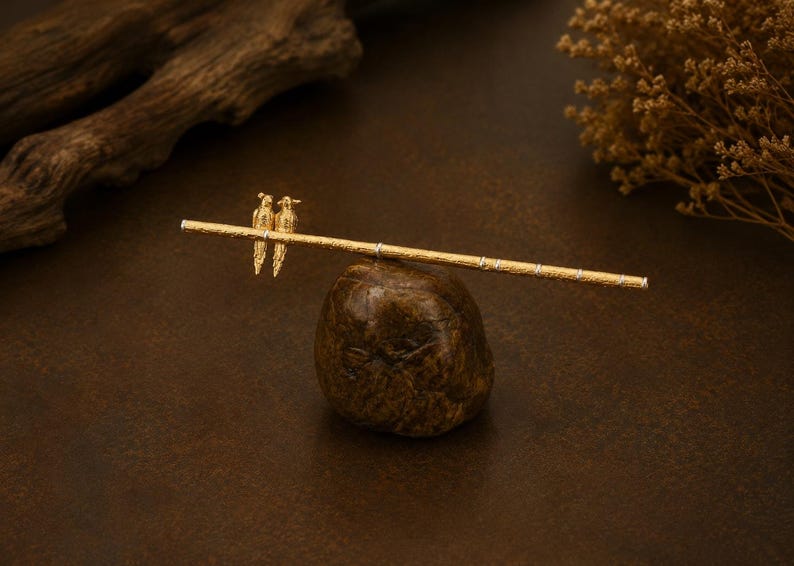 Puede incluir: Un palillo para el cabello de tono dorado con un eje texturizado similar al bamb&uacute; y dos peque&ntilde;as figuras de aves detalladas posadas en un lado. El accesorio tiene un acabado met&aacute;lico con sutiles bandas plateadas a lo largo de la varilla.