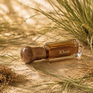 May include: A rectangular glass perfume bottle with a dark amber liquid inside and a rounded, textured brown wooden cap. The word Khus is printed in white on the front of the glass bottle. The bottle is surrounded by green grass blades.