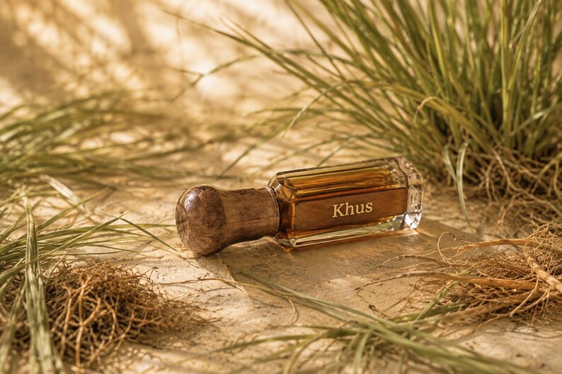 May include: A rectangular glass perfume bottle with a dark amber liquid inside and a rounded, textured brown wooden cap. The word Khus is printed in white on the front of the glass bottle. The bottle is surrounded by green grass blades.