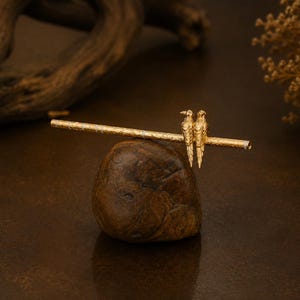 Puede incluir: Un palillo para el cabello de tono dorado con un eje cil&iacute;ndrico texturizado adornado con dos peque&ntilde;as figuras de aves detalladas posadas una al lado de la otra. La superficie met&aacute;lica muestra un patr&oacute;n fino e intrincado a lo largo de su longitud.