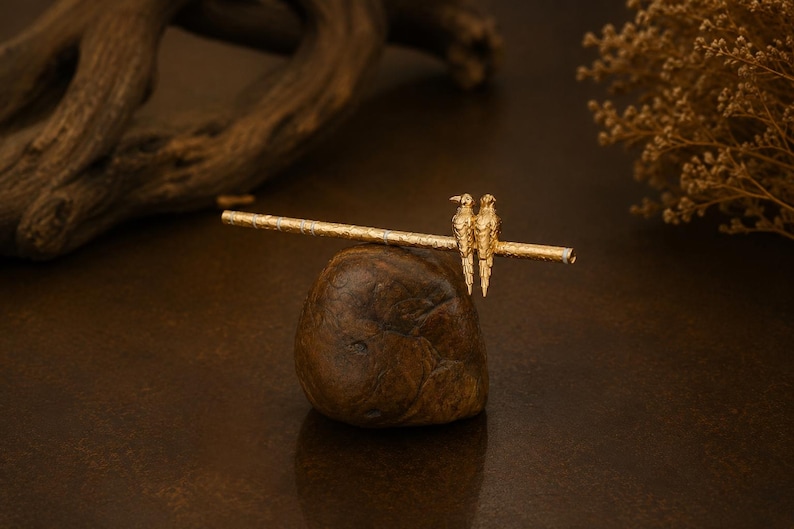 Puede incluir: Un palillo para el cabello de tono dorado con un eje cil&iacute;ndrico texturizado adornado con dos peque&ntilde;as figuras de aves detalladas posadas una al lado de la otra. La superficie met&aacute;lica muestra un patr&oacute;n fino e intrincado a lo largo de su longitud.