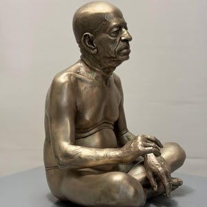 May include: A metallic bronze-toned sculpture of an elderly person with a shaved head, depicted in a seated cross-legged position. The figure has detailed facial features, visible skin texture, and is shown from a side profile view.