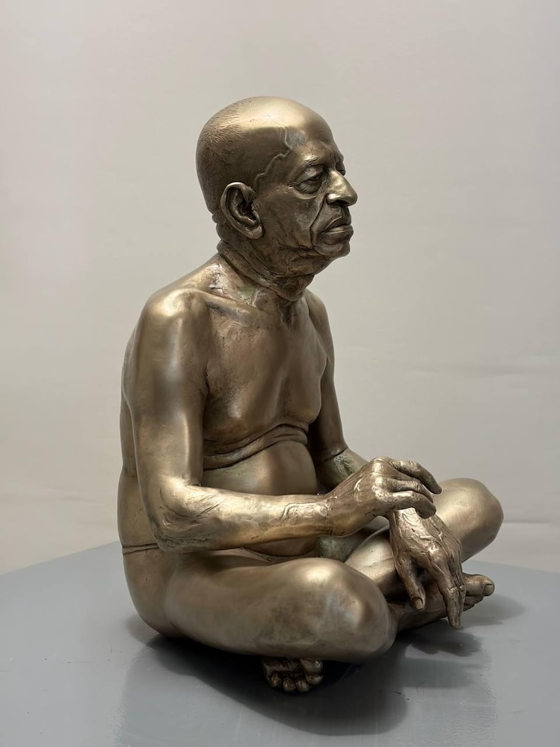 May include: A metallic bronze-toned sculpture of an elderly person with a shaved head, depicted in a seated cross-legged position. The figure has detailed facial features, visible skin texture, and is shown from a side profile view.