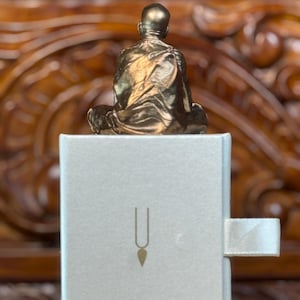 May include: A small, gold-colored statue of a seated figure atop a white box. The box features a simple design with a vertical line and a teardrop shape. The statue is detailed, with visible folds in the clothing.