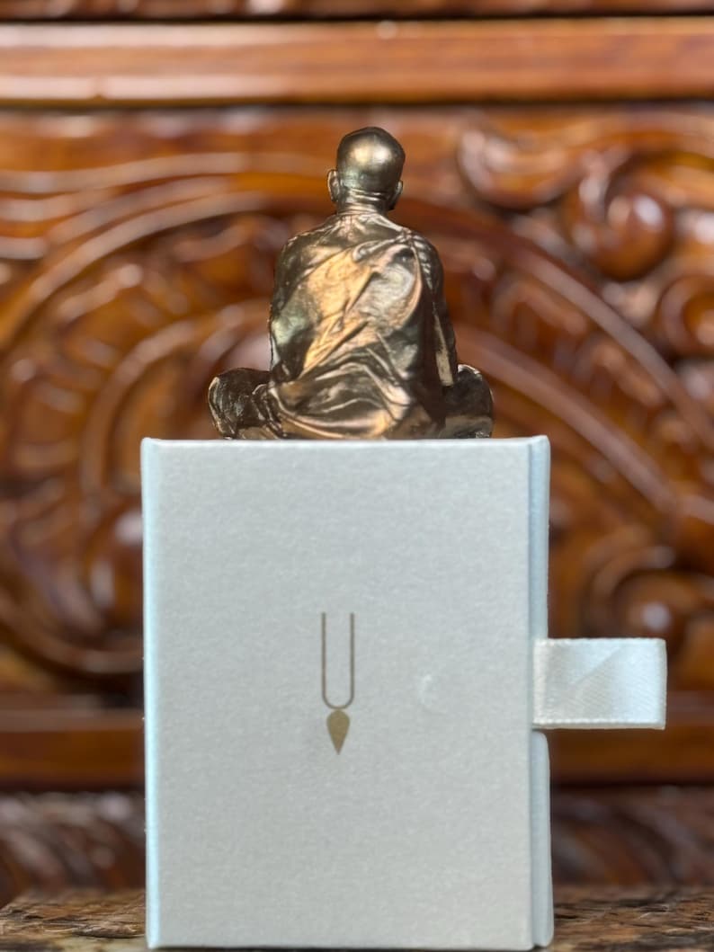 May include: A small, gold-colored statue of a seated figure atop a white box. The box features a simple design with a vertical line and a teardrop shape. The statue is detailed, with visible folds in the clothing.