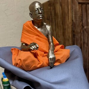 May include: A metallic bronze-toned figurine of a seated person with a shaved head, wearing a bright orange fabric robe draped over the shoulders and legs. The figure is depicted in a meditative posture with one hand resting on the lap.