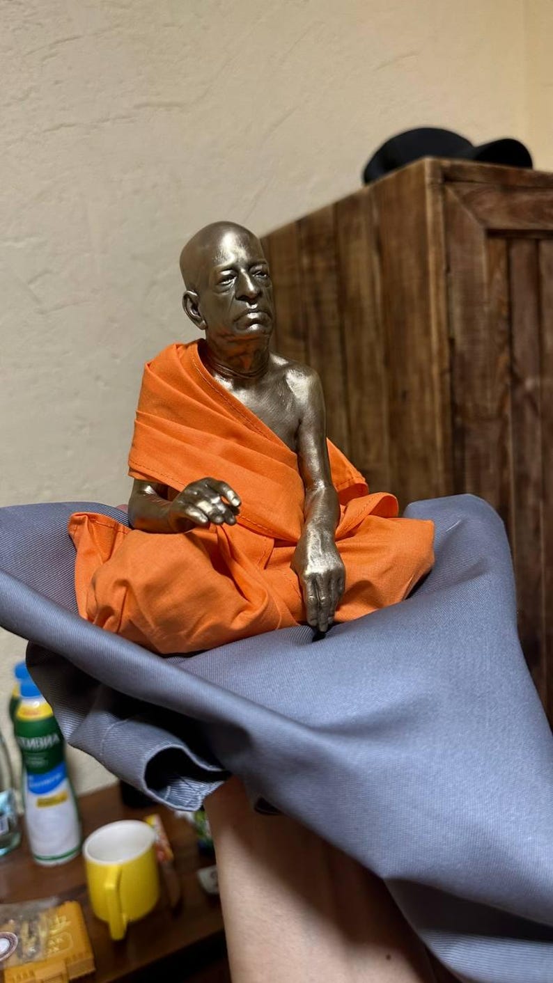 May include: A metallic bronze-toned figurine of a seated person with a shaved head, wearing a bright orange fabric robe draped over the shoulders and legs. The figure is depicted in a meditative posture with one hand resting on the lap.