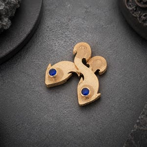 May include: A gold-toned brooch shaped like two stylized fish swimming in opposite directions. The fish feature a textured, matte surface finish and each has a single, round, deep blue gemstone set as an eye. The design is symmetrical and compact.