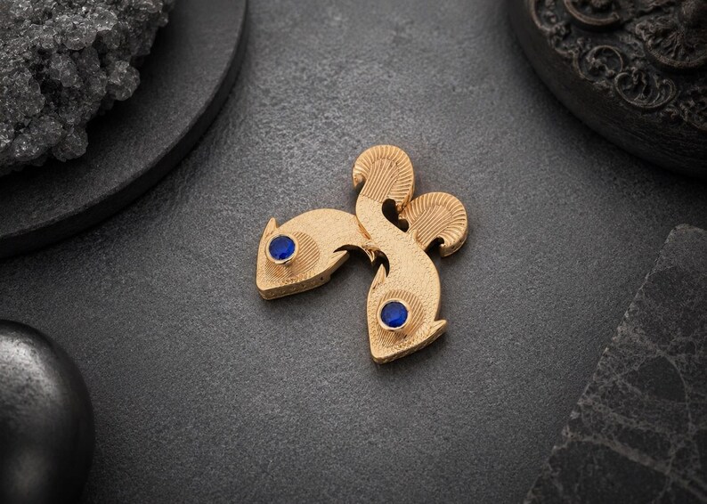 May include: A gold-toned brooch shaped like two stylized fish swimming in opposite directions. The fish feature a textured, matte surface finish and each has a single, round, deep blue gemstone set as an eye. The design is symmetrical and compact.