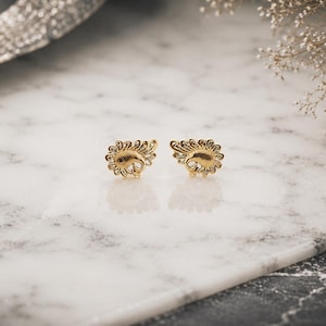 May include: A pair of gold stud earrings featuring a detailed paisley or teardrop shape with intricate swirling patterns and a textured finish. The earrings are symmetrical, showcasing a polished metallic surface that reflects light across the design.