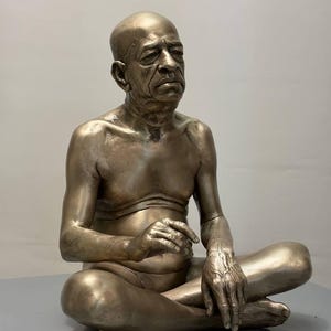 May include: A metallic bronze-toned sculpture of an elderly person with a shaved head, seated in a cross-legged position. The figure has a serious facial expression, visible skin texture, and is depicted with a bare chest and arms resting on legs.