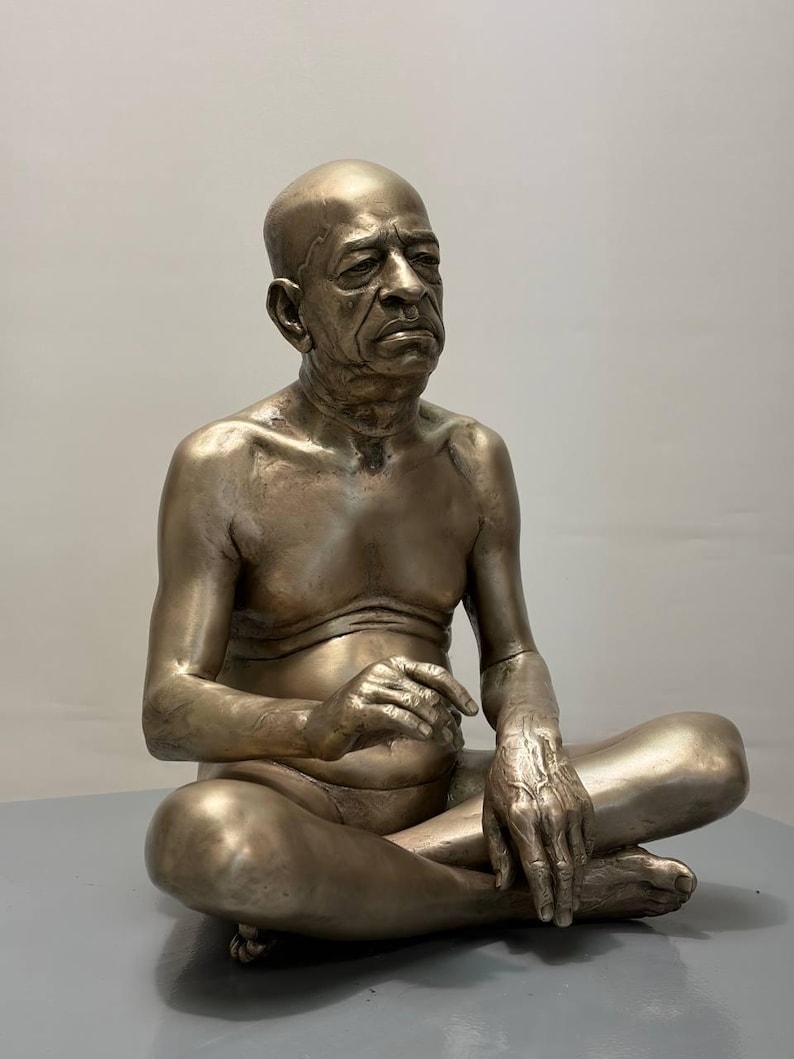 May include: A metallic bronze-toned sculpture of an elderly person with a shaved head, seated in a cross-legged position. The figure has a serious facial expression, visible skin texture, and is depicted with a bare chest and arms resting on legs.