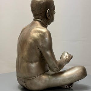 May include: A metallic bronze-toned sculpture of a seated figure in a cross-legged position. The figure has a bald head, visible skin texture, and is depicted in profile, facing right with hands positioned forward in a resting gesture.