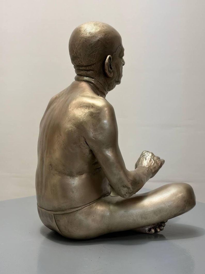 May include: A metallic bronze-toned sculpture of a seated figure in a cross-legged position. The figure has a bald head, visible skin texture, and is depicted in profile, facing right with hands positioned forward in a resting gesture.