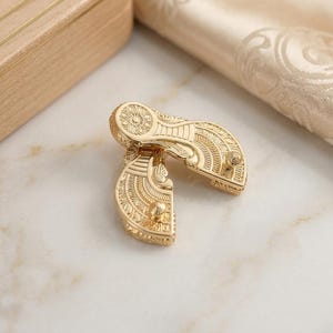 May include: A pair of gold-toned metal fasteners featuring an intricate, embossed fan-shaped design with fine linear and circular patterns. Each piece has a small, protruding stud on the surface, showcasing a polished, reflective metallic finish.