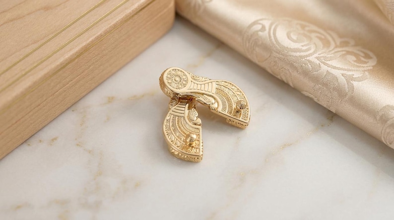 May include: A pair of gold-toned metal fasteners featuring an intricate, embossed fan-shaped design with fine linear and circular patterns. Each piece has a small, protruding stud on the surface, showcasing a polished, reflective metallic finish.