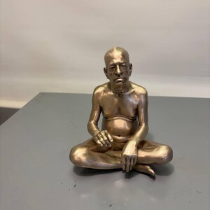 May include: A metallic bronze-colored figurine of a seated person with a shaved head, depicted in a cross-legged position. The figure has a bare chest, a visible navel, and arms resting on the legs, with a serious facial expression.
