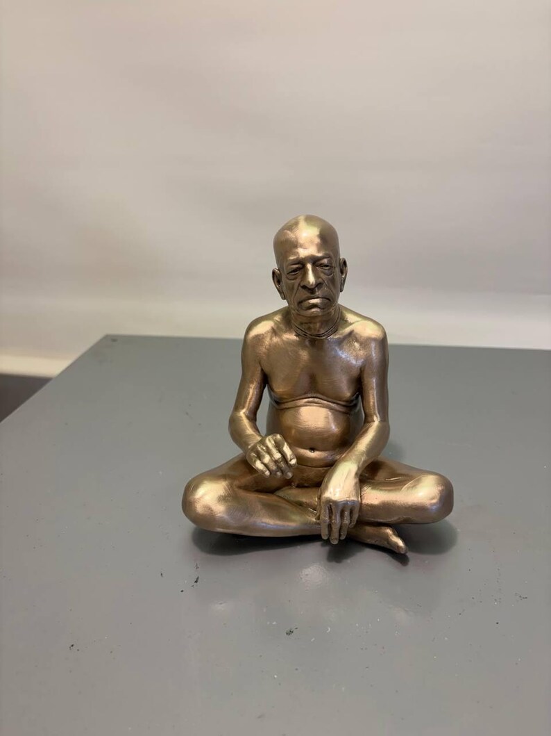 May include: A metallic bronze-colored figurine of a seated person with a shaved head, depicted in a cross-legged position. The figure has a bare chest, a visible navel, and arms resting on the legs, with a serious facial expression.