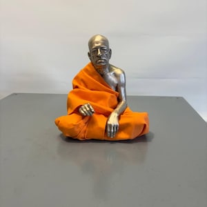 May include: A metallic silver-toned figurine of a bald individual with a serious expression, seated in a cross-legged position. The figure is draped in a vibrant orange fabric garment that covers the lower body and one shoulder, leaving the arm exposed.