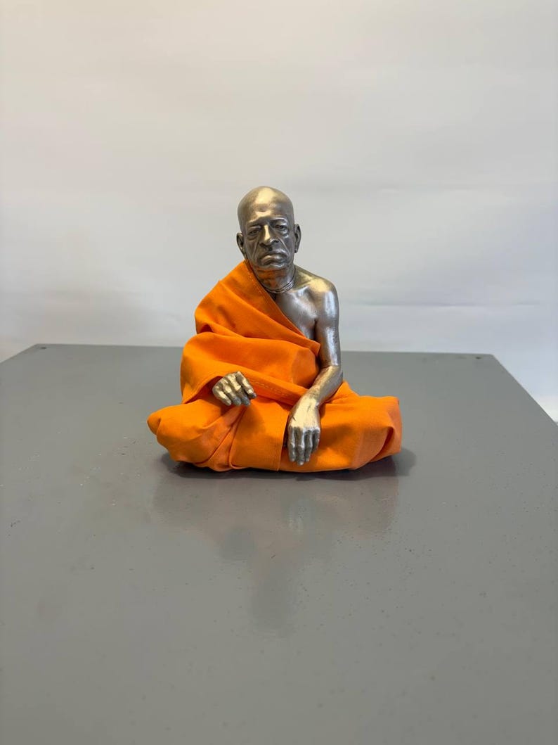 May include: A metallic silver-toned figurine of a bald individual with a serious expression, seated in a cross-legged position. The figure is draped in a vibrant orange fabric garment that covers the lower body and one shoulder, leaving the arm exposed.