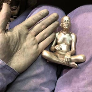 May include: A small, gold-colored statue of a seated figure in a meditative pose. The statue is detailed with visible facial features and is held near a hand covered in a similar gold-colored substance. The figure is seated with legs crossed.