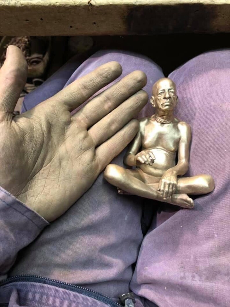 May include: A small, gold-colored statue of a seated figure in a meditative pose. The statue is detailed with visible facial features and is held near a hand covered in a similar gold-colored substance. The figure is seated with legs crossed.