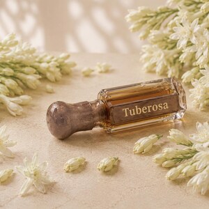 May include: A small, rectangular glass perfume bottle with a metallic, rounded cap and amber-colored liquid inside. The word Tuberosa is printed in gold lettering on the front of the bottle, which is surrounded by clusters of white tuberose flowers.