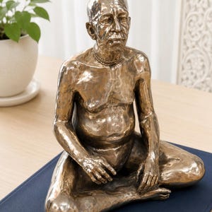 May include: A metallic bronze-toned sculpture of a seated person with a shaved head, depicted in a meditative pose with legs crossed. The figure features a textured surface, a prominent chest, and a beaded necklace around the neck.
