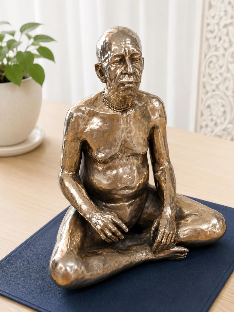 May include: A metallic bronze-toned sculpture of a seated person with a shaved head, depicted in a meditative pose with legs crossed. The figure features a textured surface, a prominent chest, and a beaded necklace around the neck.