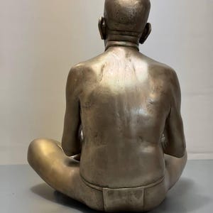 May include: A metallic bronze-toned sculpture of a person viewed from behind. The figure is seated in a cross-legged position with a shaved head and a simple loincloth. The sculpture features a smooth, reflective surface with visible muscle definition.