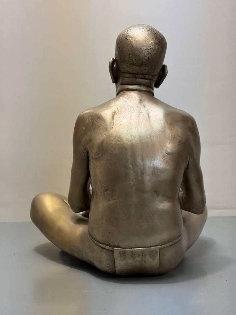 May include: A metallic bronze-toned sculpture of a person viewed from behind. The figure is seated in a cross-legged position with a shaved head and a simple loincloth. The sculpture features a smooth, reflective surface with visible muscle definition.