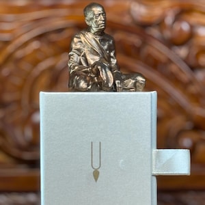 May include: A bronze-colored statue of a seated figure atop a light gray box. The box has a gold-colored symbol on the front and a white ribbon pull. The background is a wooden surface.