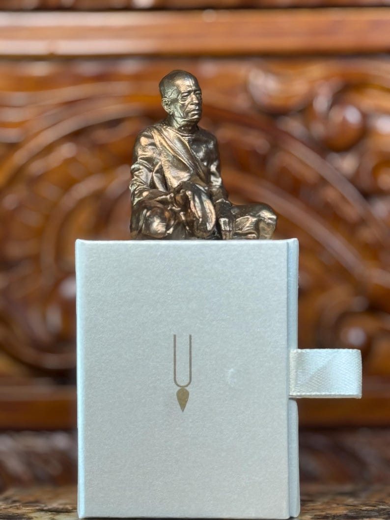May include: A bronze-colored statue of a seated figure atop a light gray box. The box has a gold-colored symbol on the front and a white ribbon pull. The background is a wooden surface.