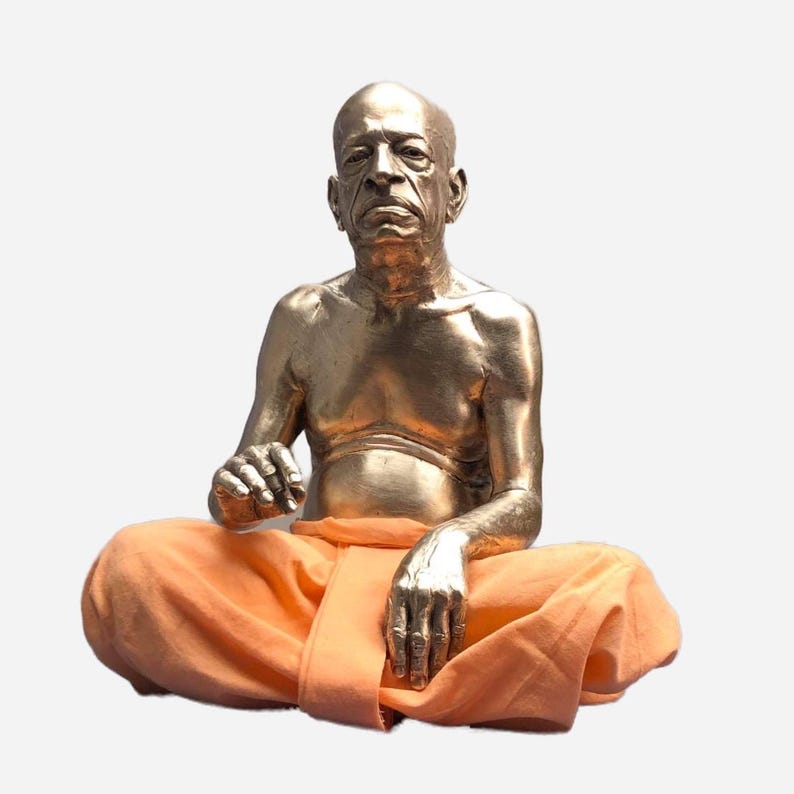 May include: A metallic gold-toned sculpture of a seated figure with a shaved head, depicted in a meditative pose. The figure has a bare chest and arms, and is wearing bright orange fabric cloth draped around the lower body and legs.