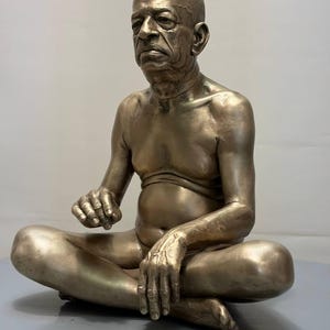 May include: A metallic bronze-toned sculpture of a seated person with a shaved head, depicted in a cross-legged position. The figure has a serious facial expression, visible skin texture, and is shown from the waist up, resting hands on legs.