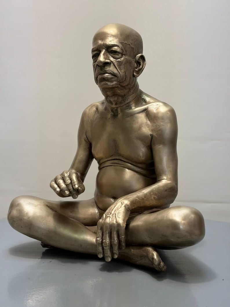 May include: A metallic bronze-toned sculpture of a seated person with a shaved head, depicted in a cross-legged position. The figure has a serious facial expression, visible skin texture, and is shown from the waist up, resting hands on legs.